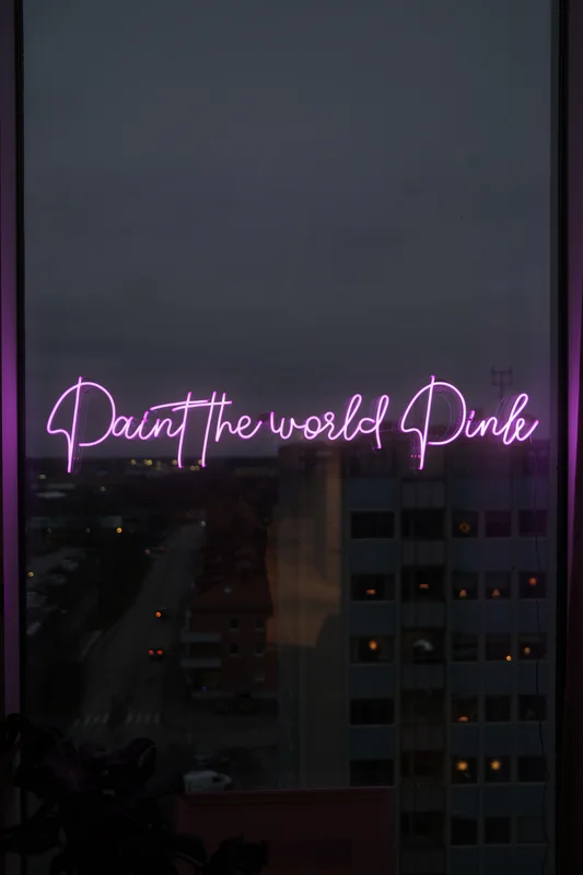 Neon pink cursive text “Paint the world Pink” over a nighttime city street and building windows.