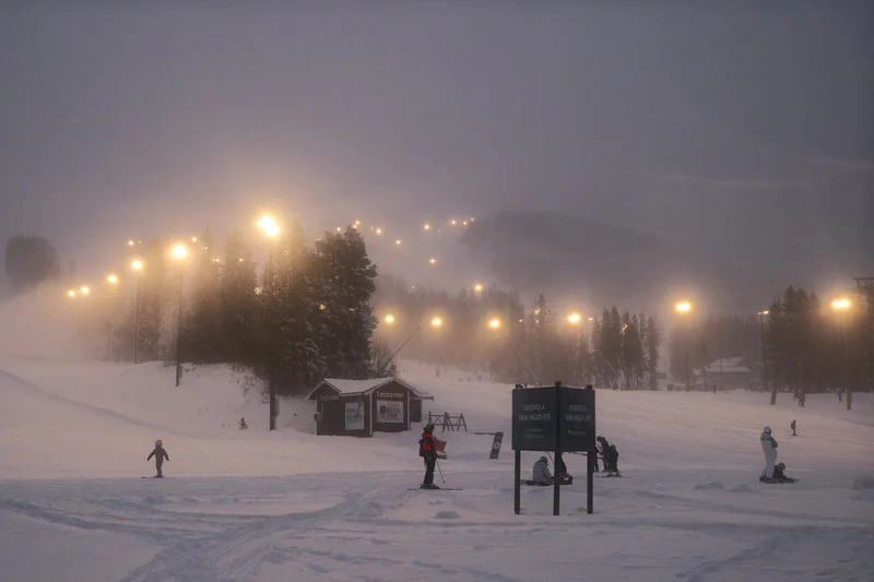 Snowy ski area at dusk with streetlights, foggy trees, and people on skis near signboards.