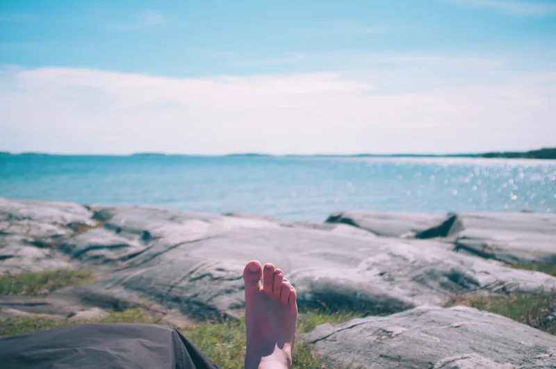 Bare feet on a rocky beach as waves break in the distance under a cloudy sky.