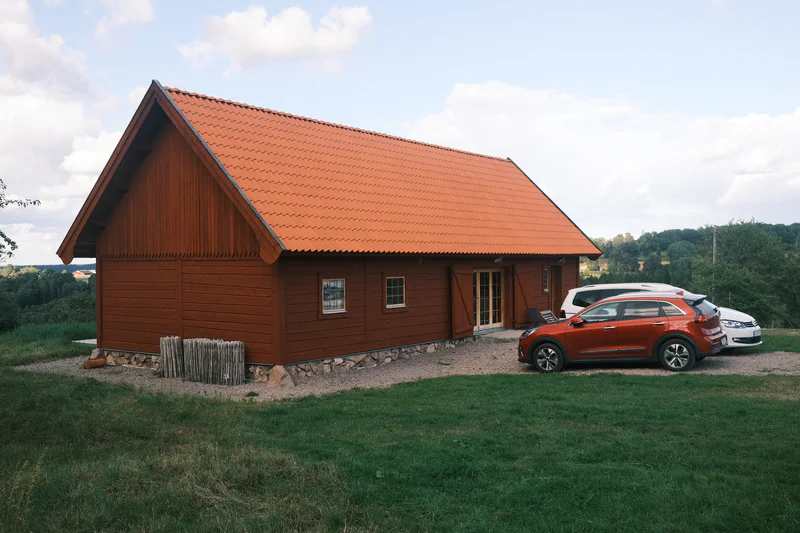 Wooden cabin with a red tile roof, a gravel driveway, and two parked cars in a grassy field.