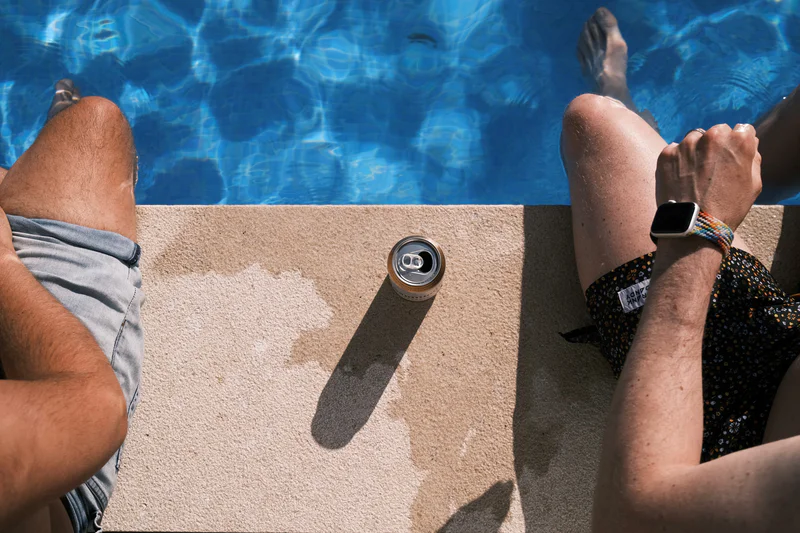 Two people lounge by a swimming pool, feet on a concrete edge next to a drink can.