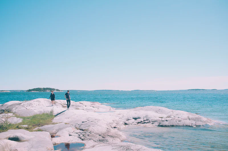 Two people stand on pale rock ledges by a turquoise sea under a clear blue sky.