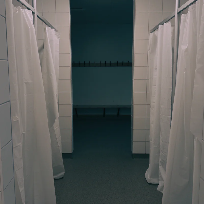 Two open shower stalls with white curtains separated by tiled walls, viewed from inside a locker room.