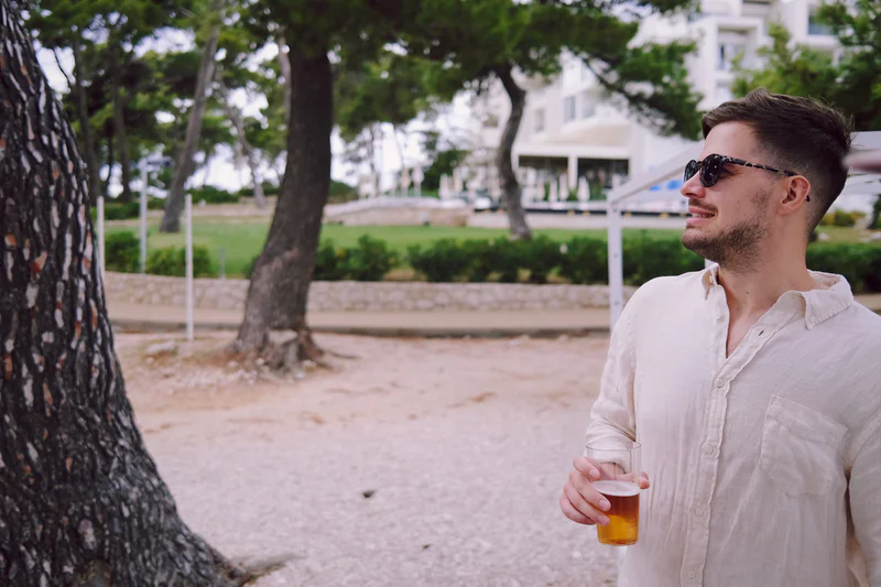 Man in a white shirt wearing sunglasses holds a yellow drink outdoors near trees and a building.