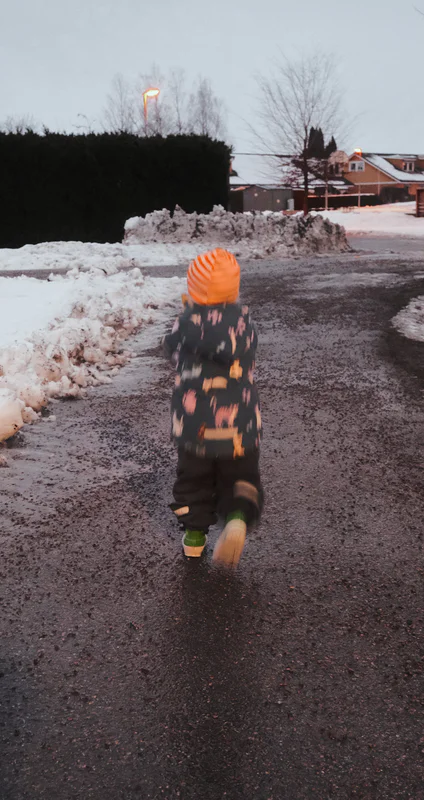 Child in an orange hat and puffy coat walks on a snowy road beside plowed snowbanks and streetlights.