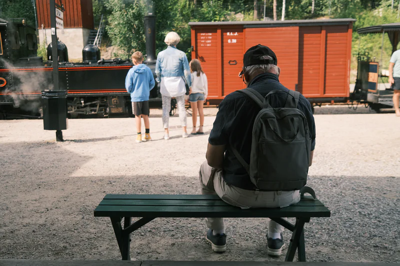 Older man with backpack sits on a green bench as children and a train pass nearby, with an orange train car in view.