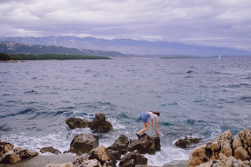 Person crouches on rocky shore above choppy ocean water with mountains and cloudy sky in the distance.