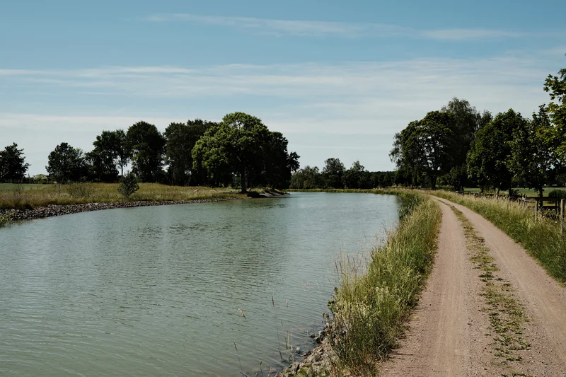 Calm canal with a dirt path along the bank, trees lining both sides under a partly cloudy sky.